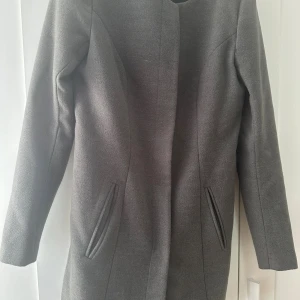 Grå klassisk kappa med fickor - Snygg grå kappa med minimalistisk design och raka linjer. Jackan har två diskreta fickor framtill och är helfodrad. Perfekt för dig som gillar stilrena plagg med enkel look. Långärmad och utan synliga knappar för en clean finish.