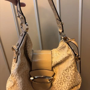 Beige axelväska från Guess - Snygg beige axelväska från Guess med klassiskt G-mönster över hela väskan. 