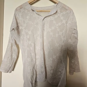 Vit broderad henley med paljetter - Säljer en vit henley-skjorta med broderade blommor och små paljettdetaljer. Skjortan är i ett lätt och transparent material, har trekvartsärm och knappar framtill. Perfekt för dig som gillar romantiska och unika plagg med extra detaljer.