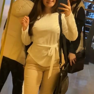 Vit ribbad långärmad topp med knyt - Säljer en vit långärmad topp med ribbad struktur och avtagbart knytband i midjan. Toppen är offshoulder på ena axeln och är figurnära utan att vara tight. Endast använd 1 gång