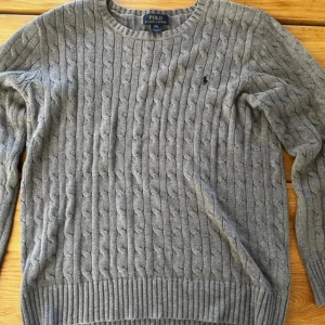 Grå kabelstickad tröja Polo Ralph Lauren - Kabelstickad grå tröja från Polo Ralph Lauren med klassisk rund halsringning och broderad logga på bröstet. Tröjan har ribbade muddar och är långärmad, perfekt för lager-på-lager-stil under kyliga dagar. Dess storlek är xl, men passar som en m.