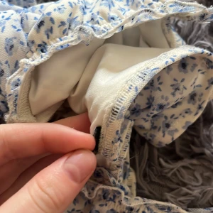 Blommig kortklänning med knuten rosett som ej går att ta av - Supersöt långklänning i vitt med blått blommönster och smala axelband. Klänningen har rynkade detaljer längs sidorna och en fin knytning vid bysten. Materialet känns som polyester med ett mjukt innerfoder. Perfekt för sommarens alla tillfällen! Den är i bra skick bara att den är ett par år gammal