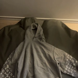 Grå Louis Vuitton hoodie med mönstrade ärmar - Säljer en grå hoodie från Louis Vuitton i storlek S, tillverkad i Italien. Tröjan har huva med snörning och coola mönstrade ärmar med LV-loggor i vitt. Perfekt för dig som vill sticka ut med en lyxig streetstyle-look.