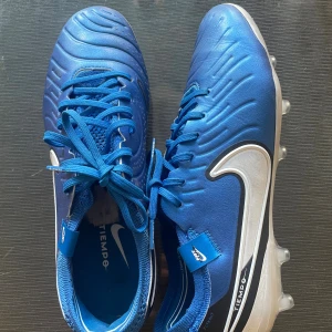 Nike Tiempo blå fotbollsskor - Säljer ett par blå Nike Tiempo fotbollsskor (Elite) med vit sula och stora vita swoosh-loggor på sidorna. Skorna har snörning, rund tå och är tillverkade i syntetmaterial med detaljer i mesh och textil. Perfekta för dig som vill sticka ut på planen.