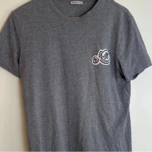 Grå Moncler t-shirt med patch - Snygg grå t-shirt från Moncler i mjuk bomull. Klassisk rund halsringning och korta ärmar. På bröstet sitter en broderad Moncler-patch i vitt och blått med röda detaljer. Perfekt för en clean och stilren look.