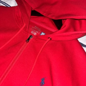 Röd hoodie från Polo Ralph Lauren - Riktigt snygg röd hoodie från Polo Ralph Lauren med dragkedja och huva. Klassisk blå broderad logga på bröstet och praktiska fickor framtill. Perfekt för dig som vill ha en stilren och bekväm tröja med streetkänsla. Priset kan diskuteras!! 