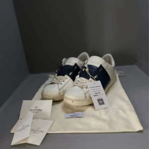 Valentino Garavani Open sneakers vit/blå - Snygga Valentino Garavani Open sneakers i vitt skinn med bred mörkblå rem över mitten. Klassisk rund tå, platt sula och coola nitar på hälen. Skorna har snörning och kommer med originalpåse och äkthetskort. Perfekta för dig som gillar exklusiva streetwear-vibbar. KAN BYTAS MOT PHILIPPE MODELL