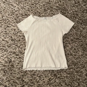 Vit ribbad topp från Lindex - Säljer en vit ribbad topp från Lindex med korta ärmar och lätt volangkant vid halsringning, ärmslut och nederkant. Toppen har en enkel, stilren look som passar perfekt till jeans eller kjol.