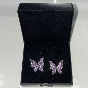 Glittriga lila fjärilsörhängen - Supersöta örhängen i form av fjärilar, täckta med lila glittrande stenar. De har en silverfärgad bas. Perfekta för dig som vill ha något unikt och färgglatt som sticker ut. Ej riktigt silver. 