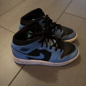 Nike Air Jordan 1 Mid blå/svart - Nike Air Jordan 1 Mid sneakers i blått och svart med klassisk snörning och rund tå. Snygg kontrast mellan den ljusblå ovandelen och svarta detaljer. Yttersulan är blå och har tydlig Nike-logga. Perfekt för dig som gillar streetwear och ikoniska sneakers.