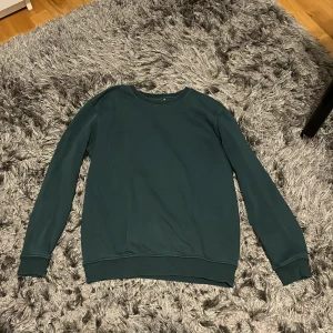 Mörkgrön sweatshirt från Kappahl - En stilren mörkgrön sweatshirt från Kappahl i storlek 146/152. Tröjan har rund halsringning, långa ärmar och ribbade muddar vid ärmslut och nederkant. Perfekt för dig som gillar en enkel och clean look.