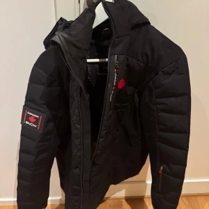 Svart pufferjacka från Canada Snow - Säljer en svart pufferjacka från Canada Snow med huva och röda detaljer. Jackan har dragkedja framtill, logga på bröstet och ärmen samt praktiska fickor med dragkedja. Perfekt för kalla dagar och snygg streetstil.