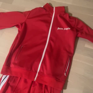 Palm Angels röda träningsbyxor - Röda träningsbyxor från Palm Angels med vita sidoränder och logga på höften. Byxorna har elastisk midja och dragkedja vid benslut. Snygg sportig look med raka ben och klassiska detaljer. Perfekta för dig som gillar streetwear.