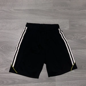 Svarta träningsshorts från D-Way - Svarta träningsshorts från D-Way med vita ränder längs sidorna och tunna gula detaljer. Shortsen har elastisk midja och är gjorda i ett lätt och svalt polyestermaterial, perfekta för sport eller gym. Klassisk sportig look med loose passform.
