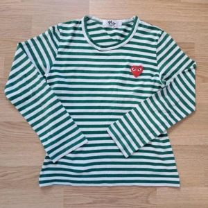 Randig långärmad tröja Comme des Garçons - Grön- och vitrandig långärmad tröja från Comme des Garçons Play i bomull. Klassisk passform med rund hals och ikoniskt rött hjärta med ögon broderat på bröstet. Perfekt för dig som gillar streetstyle och vill sticka ut med en cool detalj.