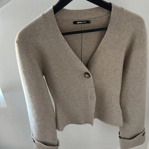 Beige kofta från Gina Tricot - Säljer en stilren beige kofta från Gina Tricot med v-ringning och knappar framtill. Koftan har långa ärmar och är tillverkad i ett mjukt stickat material. Perfekt att slänga över en topp för en chill look.