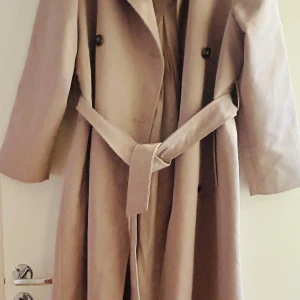 Beige trenchcoat med bälte - Stilren beige trenchcoat med dubbelknäppning och bälte i midjan. Jackan har klassisk krage, långa ärmar och raka fickor framtill. Perfekt för dig som gillar en tidlös och clean look. Materialet känns mjukt och följsamt. Storlek XS men passsar S/M använd 2 gånger som ny 