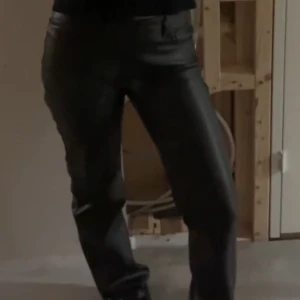 Svarta byxor i skinnimitation - Snygga svarta byxor i skinnimitation. Klassisk femficksdesign med knapp och dragkedja framtill. Perfekt för dig som vill ha en cool och stilren look med lite edge.