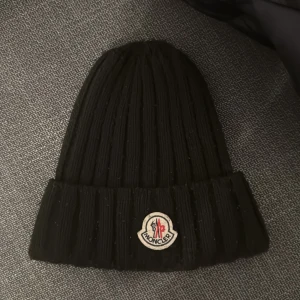 Svart ribbad Moncler mössa - Snygg svart ribbstickad mössa från Moncler med klassisk uppvikt kant och ikonisk Moncler-logga framtill. Mössan är gjord i mjukt stickat material som håller dig varm och har en clean, stilren look som funkar till alla jackor.