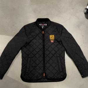 Svart quiltad jacka från Morris - Quiltad svart jacka från Morris med gul broderad logga och brittisk flagga på bröstet. Jackan har dragkedja framtill, ståkrage och två sidofickor. Snyggt rutmönster över hela jackan och detaljer i brunt vid dragkedjan. Perfekt för dig som gillar klassisk stil.