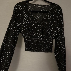 Svart prickig blus från Missguided - Säljer en svart blus från Missguided med vita prickar, croppad passform och smockad midja. Blusen har v-ringning och långa ärmar med lätt puff. Perfekt för dig som gillar trendiga detaljer och mönster.
