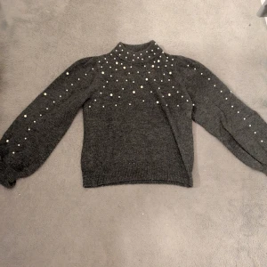 Mörkgrå stickad tröja med pärlor - Snygg mörkgrå stickad tröja med pärldetaljer över axlar och ärmar. Tröjan har hög hals och långa ärmar.