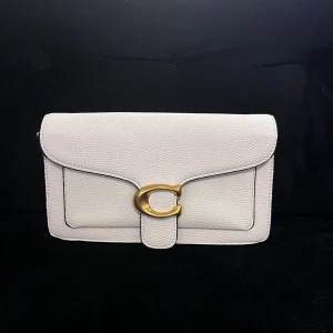 Vit axelväska från Coach i skinn - Stilren vit axelväska från Coach i strukturerat skinn med guldiga metalldetaljer och ikoniskt C-spänne framtill. Väskan har justerbar axelrem och flera fack inuti, inklusive ett med dragkedja. Perfekt för dig som gillar klassisk och clean design.