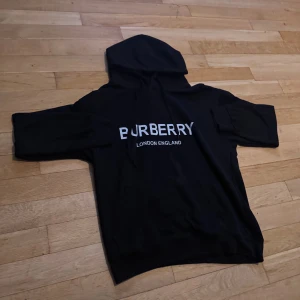 Svart Burberry hoodie med tryck - Svart hoodie från Burberry med stor vit logga och texten 'London England' på bröstet. Tröjan har huva med snörning och klassisk känguruficka framtill. Mjuk och skön, perfekt för chill dagar. Passar dig som gillar stilrena designer och exklusiva märken.