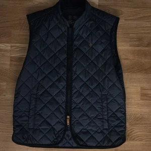 Svart quiltad väst från Morris - Stilren marinblå  quiltad väst från Morris med dragkedja framtill och diskret logga på bröstet. Västen har klassiskt rutmönster och är tillverkad i ett glansigt syntetmaterial. Perfekt för lager-på-lager och snygg till både hoodie och skjorta.