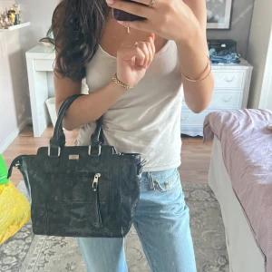 Svart handväska i skinn från Feel - Snygg svart handväska från Feel i glansigt skinn med silverfärgade metalldetaljer. Väskan har två rejäla handtag, dragkedja framtill och stilren, boxig form. Perfekt storlek för att få plats med allt du behöver och passar till många olika looks. Finns även avtagbart axelband