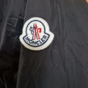 Svart vindjacka från Moncler - Svart vindjacka från Moncler med huva och coolt Moncler-märke på ärmen. Jackan har dragkedja framtill, snörning vid halsen och en snygg röd, vit och blå rand på huvan. Perfekt för dig som gillar stilren design och exklusiva märken. Säljes då den är för stor för mig. Jag väger 58 kg