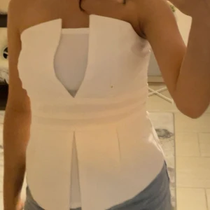 Vit tubtopp med unik v-ringning - Säljer en vit tubtopp med cool v-ringad detalj framtill och breda veckade partier vid midjan. Toppen är axelbandslös och har en stilren, modern look som passar perfekt till jeans eller kjol. Materialet känns mjukt och har en lätt struktur.