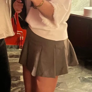 Grå plisserad minikjol - Snygg grå kortkjol med plisserad design och diskret rutigt mönster. Kjolen har en klassisk A-linjeform och sitter högt i midjan. Perfekt för dig som gillar en trendig och stilren look.