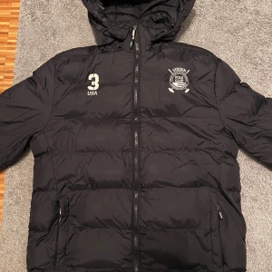 Svart pufferjacka Vinson Polo Club - Svart pufferjacka från Vinson Polo Club med huva och dragkedja. Jackan har vita tryckdetaljer på bröstet, två sidofickor med dragkedja och quiltad design. Perfekt för kalla dagar och har en sportig look med USA-tryck och klubbmärke. Pris går att diskutera vid snabb affär!!