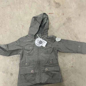 Grå rutig vindjacka från Outburst - Snygg grå vindjacka från Outburst med rutigt mönster, huva och fyra fickor med knappar framtill. Jackan har dragkedja och coolt märke på ärmen. Perfekt för dig som gillar street och funktion i ett.