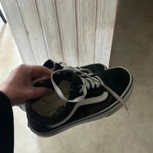Svarta Vans Old Skool sneakers - Klassiska Vans Old Skool sneakers i svart med vit sula och vit sidostrip. Skorna har snörning och en rund tå. Ovandelen är i mocka och canvas, vilket ger en skön och cool streetstil. Perfekta för dig som gillar en tidlös och avslappnad look. Använda endast ett par gånger.