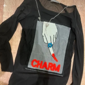 Svart mesh topp med CHARM-tryck - Cool svart mesh topp från Vince med lång ärm och ett stort tryck framtill. Trycket visar en hand med röd nagellack och blå ring, samt texten CHARM i rött. Perfekt för dig som gillar statement-plagg och unik stil.