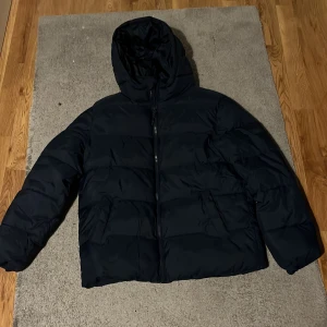 Svart pufferjacka från MNG med huva - Svart pufferjacka från MNG med huva och dragkedja framtill. Jackan har ett stilrent, quiltat utseende och en patch med texten 'THE TIME IS NOW' på ena sidan. Perfekt för kalla dagar och har en modern, loose passform varm för vintern sitter perfekt 