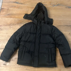 Svart dunjacka med huva från Levi's - Svart dunjacka från Levi's i storlek S, med vattenavvisande yttermaterial och varm dunisolering. Jackan har en stor huva, raka långa ärmar och två sidofickor. Perfekt för kalla dagar och har en clean, stilren look som funkar till streetwear.