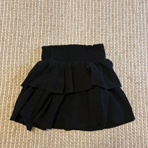 Volangkjol med smock i svart och vitt - Två korta kjolar med bred smock i midjan och dubbla volanger. En är svart och en är vit, båda i ett lätt och luftigt tyg som ger en flowig känsla. Perfekt för dig som gillar en feminin och trendig stil med extra volym.