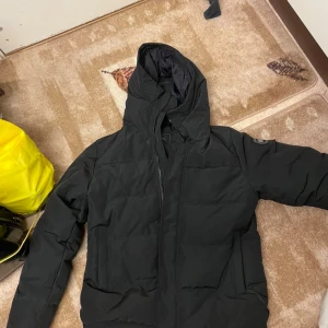  Canada Goose - Svart pufferjacka från Canada Goose med huva och diskret logotyp på ärmen. Jackan har dragkedja framtill och quiltad design som ger extra värme. Medföljer originalpåse och tags. Perfekt för kalla dagar och har en snygg, modern look.jackan e helt ny