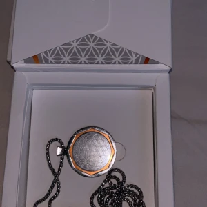 Amezcua Chi Pendant 4 halsband Energi halsband  - Unikt halsband från Amezcua, Chi Pendant 4, med rund medaljong i silverfärg och kopparfärgade detaljer. Hängs på en grov kedja och har ett geometriskt blommönster (livets blomma) på framsidan. Perfekt för dig som gillar stilrena och spirituella accessoarer.Den levereras med en katalog och en tygpåse till halsbandet.   Det är rostbeständigt, håller i 30 år och kan bäras från 12 års ålder. Om du vill veta mer om detta halsband kan du söka på Google.