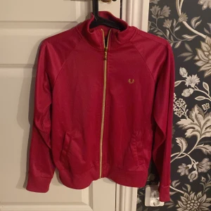 Rödrosa track jacket från Fred Perry - Säljer en snygg rödrosa track jacket från Fred Perry med guldfärgad dragkedja och logga på bröstet. Jackan har ribbade muddar, ståkrage och två fickor fram. Perfekt för dig som gillar sportig och retro stil. Helt oanvänd
