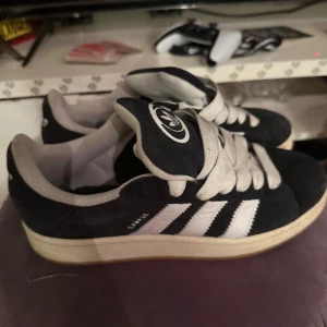 Adidas Campus svarta sneakers mocka - Svarta Adidas Campus sneakers i mocka med vita detaljer och klassiska tre ränder på sidorna. Tjocka vita skosnören och vit sula. Ikonisk Adidas-logga på plösen. Perfekta för en streetwear-look.