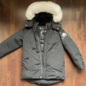Svart dunjacka Moose Knuckles storlek JR/L 152-158 - Säljer en svart dunjacka från Moose Knuckles med stor vit päls runt luvan. Jackan är använda 1 gg, perfekt skick! Nypris  7695:-, säljes flr 3500:-