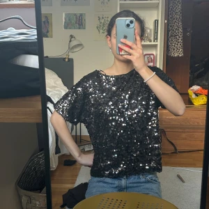 Svart paljettblus med kort ärm - Säljer en svart blus täckt med glittriga paljetter, perfekt för nyår. Blusen har rund hals och korta ärmar, och är lite loose i passformen. Har ingen bild bak då den ser lika dan ut som framsidan.