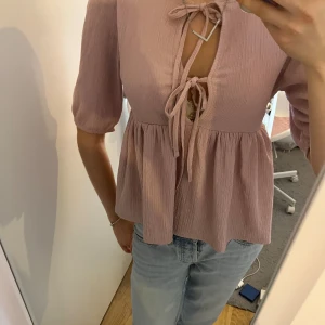 Ljusrosa blus - Puffiga korta ärmar och två knyt framtill. Blusen har en lätt rynkad, flowy passform och är gjord i ett mjukt, ribbat material. 