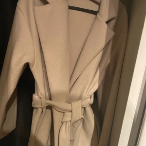 Beige kappa med bälte och bred krage - Stilren beige kappa med bred krage och knytskärp i midjan. Jackan har en klassisk omlottmodell och är tillverkad i ett mjukt material som ger en clean och modern look. Perfekt för dig som gillar minimalistisk stil och vill ha en snygg jacka till höst och vinter. Pris går att diskutera!