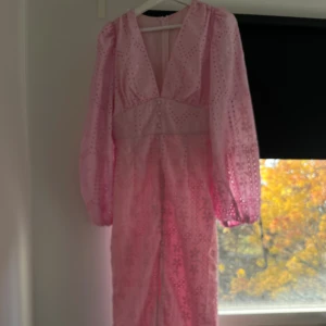 Lång rosa klänning med broderi - Säljer en lång rosa klänning med v-ringning och puffiga långärmar. Klänningen har broderade blommor, spetsdetaljer och dekorativa knappar framtill. Materialet är lätt och har ett genombrutet mönster som ger en romantisk vibe. Passformen är slim och det finns en slits framtill.