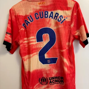 FC Barcelona Pau Cubarsí matchtröja Nike - Säljer en unik FC Barcelona fotbollströja med Pau Cubarsí och nummer 2 på ryggen. Tröjan är röd med ljusa inslag i ett unikt mönster, har korta ärmar, svarta detaljer och klubbmärke samt sponsorloggor. Perfekt för dig som älskar Barca och vill sticka ut på läktaren.